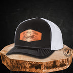 A black and white trucker hat with a brown leather patch on the front, displayed on a rustic wood slice against a dark textured background.