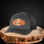 A dark charcoal gray trucker hat with a brown leather patch on the front, displayed on a rustic wood slice against a dark textured background. The patch reads 'Ol Hail Trail'.