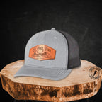 A light gray and black mesh trucker hat with a brown leather patch on the front that reads 'Ol Hail Trail' The hat is displayed on a rustic wood slice against a dark textured background.