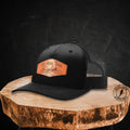 A black trucker-style cap with a curved brim and black mesh back, featuring a tan rectangular patch on the front that reads 'Ol Hail Trail' with a mountain graphic. The cap is displayed on a rustic wood slice against a dark textured background.