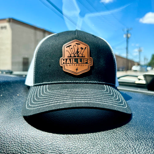 A black and white trucker hat with a brown leather patch on the front that reads 'HAIL LIFE CRAFTSMANSHIP', resting on a car dashboard with a blurred outdoor background.