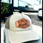 A light beige trucker hat with a mesh back and a brown leather patch on the front that reads 'Ol' Hail Trail' The hat is shown outdoors with an RV and gravel background.